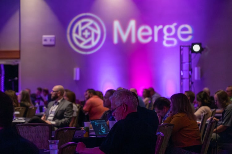 A group of people sitting in chairs in front of a wall with a logo on it Description automatically generated